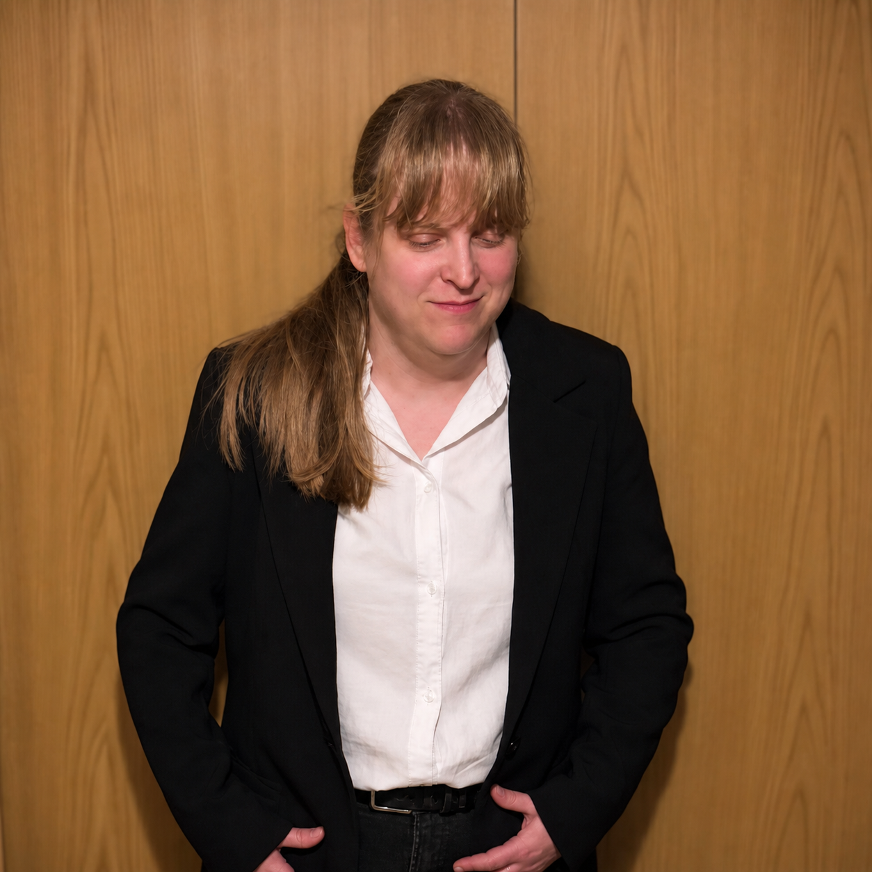A woman standing against a wooden panel background. She has light brown hair tied back in a low ponytail with bangs covering the forehead. She is wearing a white button-up shirt tucked into black pants paired with a black blazer. Her hands are resting near her waist, and she has a slight smile while looking downward. The overall setting appears professional, and the lighting is soft and even.